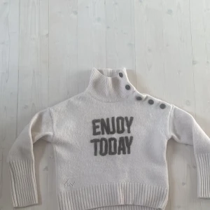 Vit stickad tröja från Zadig & Voltaire - Säljer en superfin vit stickad tröja från Zadig & Voltaire. Den har texten 'ENJOY TODAY' i grått på framsidan och coola knappar vid halsen. Tröjan är långärmad och har ribbade muddar vid ärmslut och nederkant. Perfekt för höst och vinter!