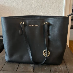 Svart axelväska från Michael Kors - Säljer en stilren svart väska från Michael Kors. Väskan är i skinn och har en elegant design med guldfärgade detaljer. Den har en rymlig insida med flera fack och en dragkedja för säker förvaring. Kommer med en dustbag