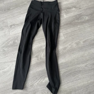 Svarta leggings från Nike - Snygga svarta leggings från Nike med Dri-FIT-teknologi för optimal komfort. Perfekta för träning med högmidjad design och en liten vit Nike-logga på benet. Tillverkade i ett stretchigt syntetmaterial som ger en tight passform. Och har en ficka på benat och en i back.