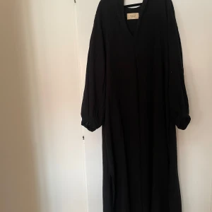 Svart klänning från A part of the art - En svart långärmad klänning med V-ringning och en lös passform. Klänningen har en slits i sidan och är gjord av ett mjukt och bekvämt tyg. Oanvänd. Nypris 2000kr. Storlek XS/S. Rökfritt hem. Skickar paket som spårbart.
