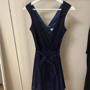 Mörkblå glittrig klänning  - Snygg mörkblå klänning från Bubbleroom med glittrigt material och omlott-design. Klänningen har en v-ringad hals och ett knytband i midjan för en smickrande passform. Perfekt för en festlig kväll! 💙Använd 1 gång