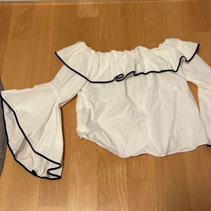 Vit offshoulder blus från Zara - Säljer en stilren vit offshoulder blus från Zara Basic Collection. Blusen har volangdetaljer och vida ärmar med svarta kanter som ger en elegant touch. Perfekt för att lyfta vilken outfit som helst!