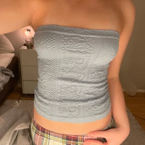 Ljusblå tubtopp med broderat mönster från orban outfitters  - Säljer en stilren ljusblå tubtopp med vackert broderat mönster. Perfekt för en trendig look. Toppen är axelbandslös och har en tight passform.
