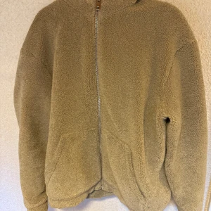 Beige teddyjacka från H&M - Mysig beige teddyjacka från H&M med dragkedja framtill och huva. Perfekt för kyliga dagar när du vill hålla dig varm och bekväm. Jackan har långa ärmar och två praktiska fickor framtill. En stilren och enkel design som passar till det mesta.