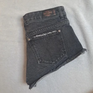 Svarta jeansshorts från Pull&Bear - Snygga svarta jeansshorts från Pull&Bear med slitna detaljer och fransiga kanter. Perfekta för en avslappnad stil. De har en klassisk femficksdesign och knappgylf.