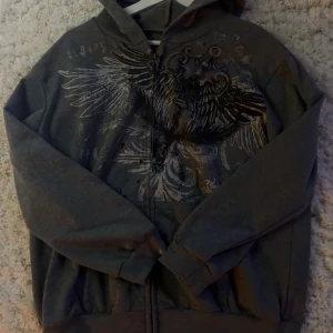 Grå hoodie med tryck - Säljer en grå hoodie med ett detaljerat tryck av vingar och mönster på framsidan. Tröjan har en dragkedja och en luva, perfekt för en avslappnad stil. Den är bekväm och passar bra till vardags.