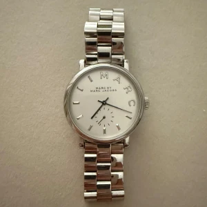 Silverfärgad klocka från Marc Jacobs - Elegant silverfärgad klocka från Marc Jacobs med vit urtavla och stilrena indexmarkeringar. Klockan har en klassisk länkarmband i metall och en subtil sekundvisare. Perfekt för en stilren look! Batteriet behövs dock bytas ut ❣️
