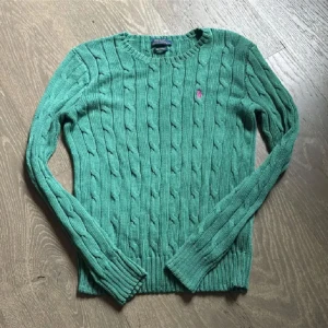 Grön stickad tröja från Ralph Lauren - Säljer en snygg grön stickad tröja från Ralph Lauren i Pima bomull. Tröjan har ett klassiskt kabelstickat mönster och en rosa logotyp på bröstet. Perfekt för kyligare dagar! Passar även S
