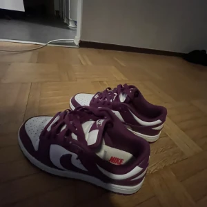 Lila och vita Nike sneakers - Snygga lila och vita Nike sneakers med klassisk design. Skorna har en vit bas med lila detaljer och snörning. Perfekta för en sportig look.