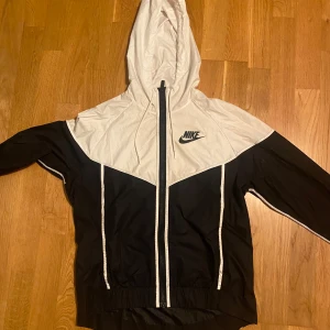 Nike vindjacka - Säljer en stilren svart och vit vindjacka från Nike med huva och dragkedja. Jackan har långa ärmar och en klassisk Nike-logga på bröstet. Perfekt för blåsiga dagar.