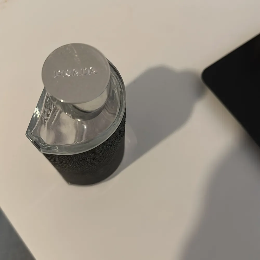 Elegant parfymflaska från Prada med en stilren design. Flaskan är genomskinlig med svarta detaljer och har en silverfärgad kork. Perfekt för den som söker en lyxig doftupplevelse.. Perfume.