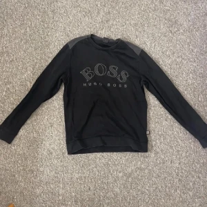 Svart tröja från Hugo Boss - Säljer en stilren svart tröja från Hugo Boss med deras logga tryckt på framsidan. Tröjan har långa ärmar och en klassisk rund halsringning. Perfekt för en avslappnad och trendig look. Den är sällsyn och svårt att hitta. Passar även storlek S.