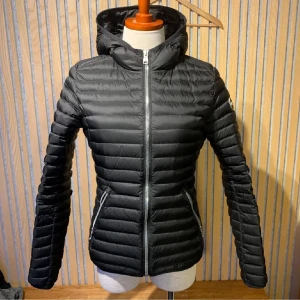 Svart dunjacka från Colmar - Snygg svart dunjacka från Colmar med huva och dragkedja framtill. Jackan har en quiltad design och två praktiska fickor med dragkedjor. Perfekt för kyligare dagar!
