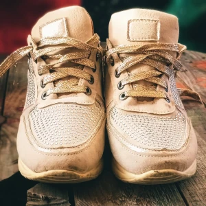 Beige/guldiga sneakers med glittriga detaljer - Snygga beige/guldiga sneakers med glittriga detaljer och gyllene snörning. Skorna har en bekväm sula och är dekorerade med små glittrande stenar på sidorna och framtill. Perfekta för att ge en touch av glamour till din outfit. Skorna är i storlek 37.