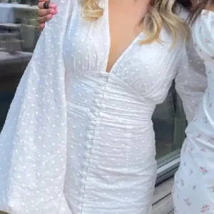 Vit somrig klänning - Säljer en söt vit klänning med prickigt mönster och v-ringad design. Klänningen har långa ärmar och knappar framtill, vilket ger en elegant touch. Perfekt för sommarens alla tillfällen! Säljer den då jag endast använt den en gång