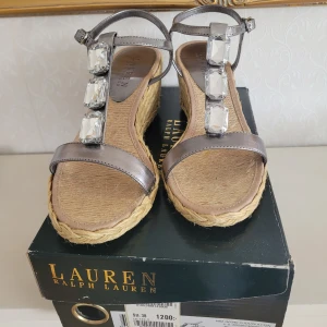 Sandaler med kilklack från Lauren Ralph Lauren - Snygga sandaler med kilklack från Lauren Ralph Lauren. De har en läder rem i silver och är dekorerade med stora, klara stenar. Sulan är i flätad jute vilket ger en somrig känsla. Perfekta för att lyfta din stil med en touch av glamour.