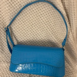 Blå axelväska i syntet - Snygg blå axelväska med krokodilpräglat mönster. Väskan har en justerbar axelrem och stängs med en magnetknapp. Perfekt för att ge en färgklick till din outfit.