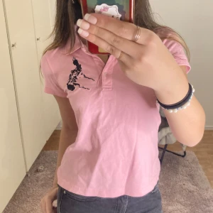 Rosa pikétröja med tryck - Säljer en rosa pikétröja med svart tryck på bröstet och ryggen. Tröjan har en klassisk krage och korta ärmar, perfekt för en avslappnad stil. Den är gjord i ett mjukt material som känns skönt mot huden.