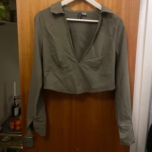Olivgrön blus - Säljer en stilren olivgrön blus. Blusen har långa ärmar och en djup v-ringning. Den är croppad och har en klassisk krage. Perfekt för en trendig look.