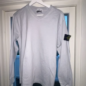 Vit/grå  långärmad tröja från Stone Island - Säljer en grå/vit långärmad tröja från Stone Island. Tröjan har en v-ringad hals och det ikoniska Stone Island-märket på ärmen. 