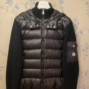 Svart Moncler Cardigan - Snygg svart cardigan från Moncler. Jackan har en dragkedja framtill och en praktisk huva. Moncler-logotypen finns på ärmen. Perfekt för kyligare dagar. Storleken är L och alla qr koder och liknande fungerar. Kommer med påse💯