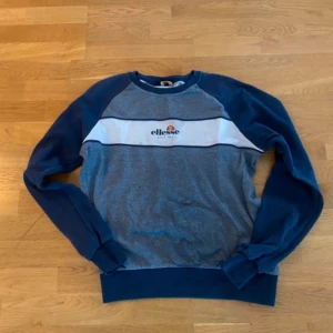 Blå och grå tröja från Ellesse - Säljer en snygg blå och grå tröja från Ellesse med en vit rand och logotyp på bröstet. Tröjan har långa ärmar och en rund halsringning. Perfekt för en avslappnad stil.