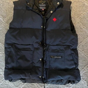 Svart dunväst från Canada Goose - Säljer en navy blue freestyle dunväst från Canada Goose med en röd lönnlövdetalj på bröstet. Västen är en äldre modell och har ett högt värde.  Västen har praktiska fickor och stängs med dragkedja och knappar. Perfekt för kyliga dagar eller nu för våren. Den är i väldigt bra skick och fungerar felfritt. Hör av er vid det minsta mvh Axel. 