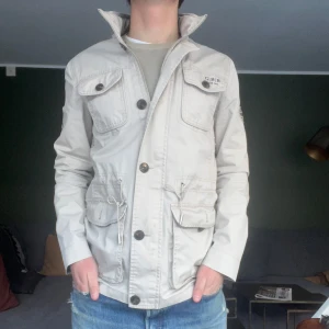 Beige fieldjacket från Henri Lloyd - Säljer en beige fieldjacka från Henri Lloyd med flera fickor och knappar. Jackan har en stilren design med broderade detaljer och hög krage. Perfekt för vår och höst.
