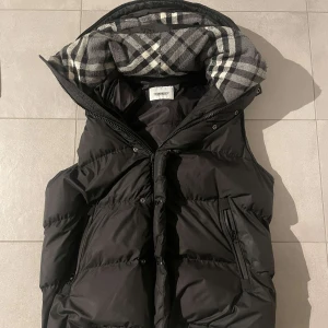 Svart dunväst från Burberry - Säljer en snygg svart dunväst från Burberry med ett klassiskt rutigt mönster. Bra skick men dragkedjan var trasig så den byttes ut, och därför står det inte ”burberry” på den. Även en knapp längst nere är borta men den märks inte. Pris kan diskuteras vid snabb affär!