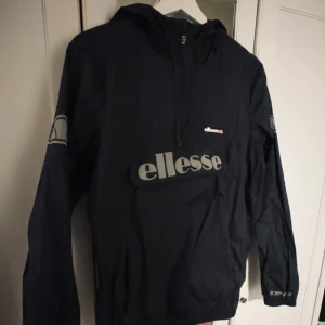 Svart vindjacka från Ellesse - Säljer en svart vindjacka från Ellesse med stor logga på bröstet och mindre loggor på ärmarna. Jackan har en huva och dragkedja framtill. Perfekt för blåsiga dagar. De gråa detaljerna som texten på magen och loggan på armen är i reflex.