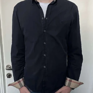 Burberry skjorta  - Snygg svart skjorta med långa ärmar och stilrena knappar. Den har en cool detalj med rutigt mönster på insidan av ärmsluten. Perfekt för en stilren look!