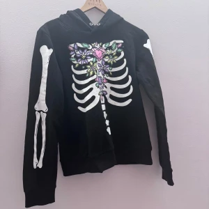 Svart hoodie med skelettmönster - Cool svart hoodie med ett unikt skelettmönster på framsidan och ärmarna. Mönstret inkluderar färgglada blommor och stenar som ger en extra touch. Perfekt för dig som vill sticka ut! Är i storlek 170