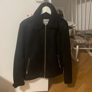 Svart jacka från Jack & Jones - Snygg svart jacka från Jack & Jones med dragkedja framtill. Jackan har en klassisk krage och är perfekt för kyligare dagar. Den har praktiska fickor med dragkedjor för säker förvaring.