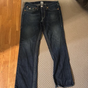 Mörkblå jeans från True Religion - Snygga mörkblå jeans från True Religion med kontrasterande vita sömmar och klassiska femficksdesign. Jeansen har en straight passform och är dekorerade med märkets ikoniska broderi på bakfickorna. Snyggt slitna på nedre bakbenen