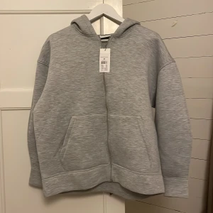Grå hoodie med dragkedja - Säljer en stilren grå hoodie med dragkedja och huva. Tröjan har långa ärmar och en praktisk ficka framtill. Perfekt för en avslappnad look. Helt ny, har lappen på och bara satte på mig den för bilden. 