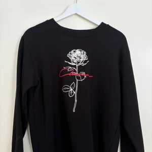 Svart tröja med rosdetalj - Snygg svart tröja med en vit ros och röd text på framsidan. Tröjan har långa ärmar och en rund halsringning. Perfekt för en stilren look.