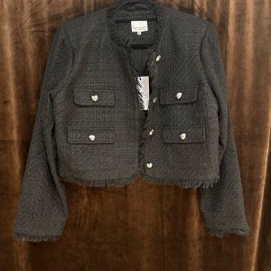 Svart kavaj från Noella - Snygg svart kavaj från Noella med fransiga kanter och hjärtformade knappar. Kavajen har två bröstfickor och en stilren design som passar perfekt för en chic look. Perfekt för att lyfta din outfit med en touch av elegans.