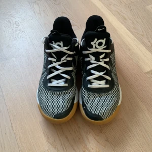 Svartvita Nike sneakers - Snygga svartvita Nike KD basketskor med ett unikt mönster och vit snörning. Skorna har en gummisula i brun färg och är perfekta för en sportig look.