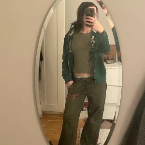 Gröna cargopants med broderi från Ed Hardy  - Snygga gröna cargopants med broderade detaljer i rött och vitt. Byxorna har en loose passform och är perfekta för en avslappnad stil. De har en lågmidjad design och coola mönster som ger en unik touch.