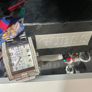 Silverfärgad klocka från Guess - Snygg silverfärgad klocka från Guess med rektangulär urtavla och glittriga detaljer runt kanterna. Pris går att diskuteras!!