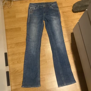 Blå bootcut jeans från wrangler - Snygga blå rock 47 jeans från Wrangler med broderade detaljer och paljetter på bakfickorna. De har en bootcut-stil och är perfekta för en avslappnad look. Jeansen har en klassisk femficksdesign och en bekväm passform.