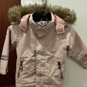 Rosa vinterjacka från Kaxs - Säljer en söt rosa vinterjacka från Kaxs i storlek 98. Jackan har en mysig fuskpäls på luvan och praktiska dragkedjor på fickorna. Perfekt för kalla vinterdagar! Lite slitage finns, men den är fortfarande i bra skick. Passar perfekt för lek och bus i snön! ❄️