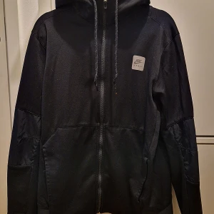 Svart huvtröja med dragkedja, Nike - Säljer en svart huvtröja / träningsjacka med dragkedja från Nike i mycket bra skick.  Snygg och stilren design och bekväm passform. Passar både till träning och vardagsbruk. Är herrstorlek man kan givetvis bäras av kvinnor också.