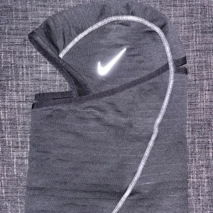Svart Nike balaclava med reflexer - Snygg och funktionell balaclava från Nike, perfekt för kalla dagar. Den är tillverkad i ett mjukt och stretchigt material med reflex sömmar och har en Nike-logga som också är reflex framtill. Passar bra för utomhusaktiviteter som löpning eller cykling under höst och vinter.