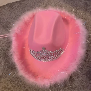Rosa cowboyhatt med tiara - Supercool rosa cowboyhatt med fluffig kant och en glittrig tiara framtill. Perfekt för fester eller när du bara vill känna dig som en prinsessa. Hatten har en justerbar rem för perfekt passform. Gör ett statement med denna unika accessoar!