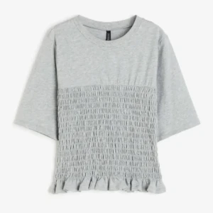 Grå topp med volangdetalj - Säljer en söt grå topp från Hm med volangdetalj längst ner. Toppen har en smockad design på framsidan och trekvartsärmar. Perfekt för en avslappnad stil eller att klä upp med accessoarer.