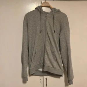 Grå ziphoodie från Jack & Jones - Säljer en stilren grå ziphoodie från Jack & Jones i mycket bra skick. Den har en dragkedja framtill och en skön huva med snören. Perfekt för både höst och vår, och passar till vardags. Storleken är normal och materialet är mjukt och bekvämt.