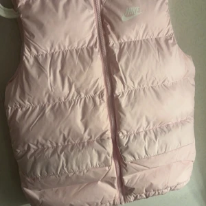Rosa dunväst från Nike - Säljer en superfin rosa dunväst från Nike. Perfekt för kyliga dagar med sin mysiga och varma design. Västen har en dragkedja framtill och är i mycket bra skick. Den är både snygg och praktisk för höst och vår.