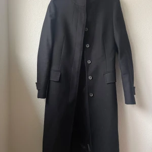 Svart kappa från Zara Woman - Säljer en elegant svart kappa från Zara Woman i storlek M. Den har en stilren design med knappar framtill och en hög krage. Perfekt för höst och vinter, och ger en sofistikerad look. Kappan är i mycket bra skick och är tillverkad i ett mjukt material som känns lyxigt.