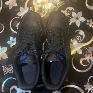 Svarta Nike Air sneakers - Säljer ett par svarta Nike Air force i skinn med blå detaljer. Skorna har klassisk snörning och en stilren design. De är i mycket bra skick och redo för nya äventyr! 
